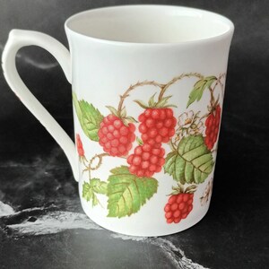 K&ouml;nnte beinhalten: Wei&szlig;e Keramiktasse mit Henkel, mit einem roten Himbeerdesign auf wei&szlig;em Hintergrund.