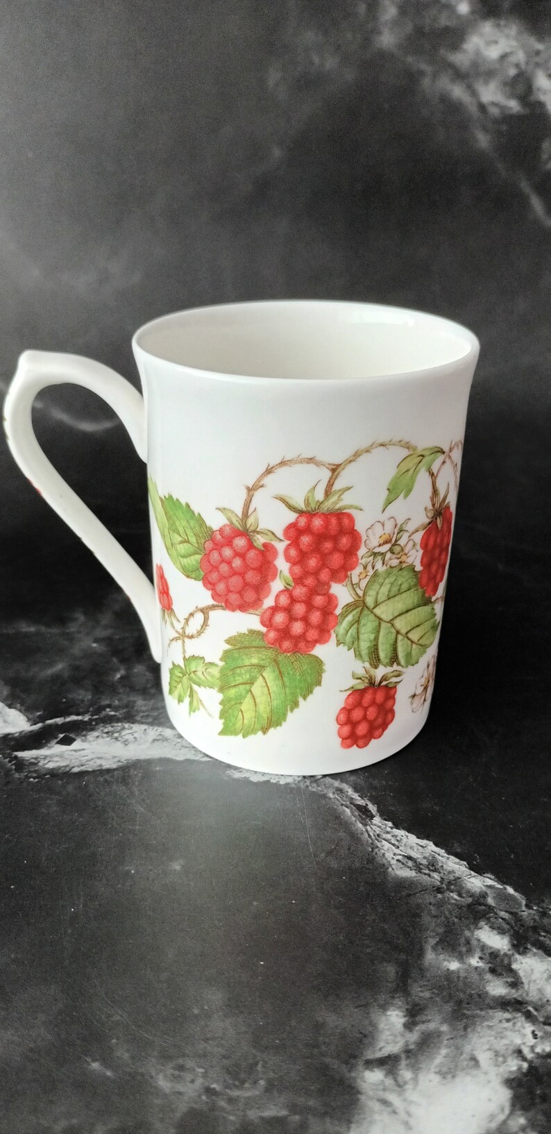 K&ouml;nnte beinhalten: Wei&szlig;e Keramiktasse mit Henkel, mit einem roten Himbeerdesign auf wei&szlig;em Hintergrund.