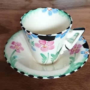 Op de afbeelding: Een vintage theekop en schotel set. De witte keramische kop en schotel zijn versierd met handgeschilderde roze en blauwe bloemmotieven, groene bladeren en zwarte stippen. De kop heeft een uniek hoekig handvat.