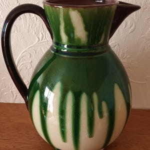 May include: A ceramic pitcher featuring a glossy finish with a cream-colored base and dark green vertical drip glaze patterns. The vessel has a rounded body, a wide pouring spout, and a dark brown curved handle attached to the side of the rim.