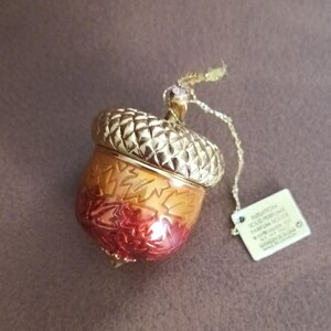 May include: A gold-toned acorn-shaped solid perfume container with a textured cap and a gradient orange-to-red base featuring embossed leaf patterns. A gold cord is attached to the top, holding a small white tag with the text Intuition Solid Perfume.