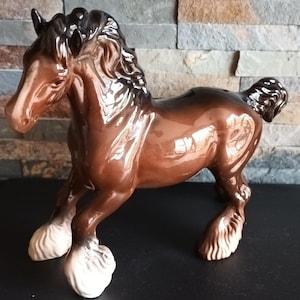 May include: A brown ceramic horse figurine in a trotting pose. The horse has a glossy finish, a dark mane and tail, and white feathered feet. The figurine has a slot on its back, suggesting it may be a piggy bank. The horse is approximately 25 cm long.