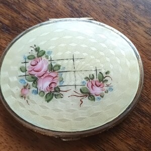 May include: A vintage, round compact with a pale yellow, textured surface. The compact features a floral design of pink roses with green leaves and blue accents. The compact has a silver-colored metal rim.