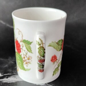 K&ouml;nnte beinhalten: Wei&szlig;e Keramiktasse mit Henkel, verziert mit einem Blumendesign mit gr&uuml;nen Bl&auml;ttern und roten Beeren.