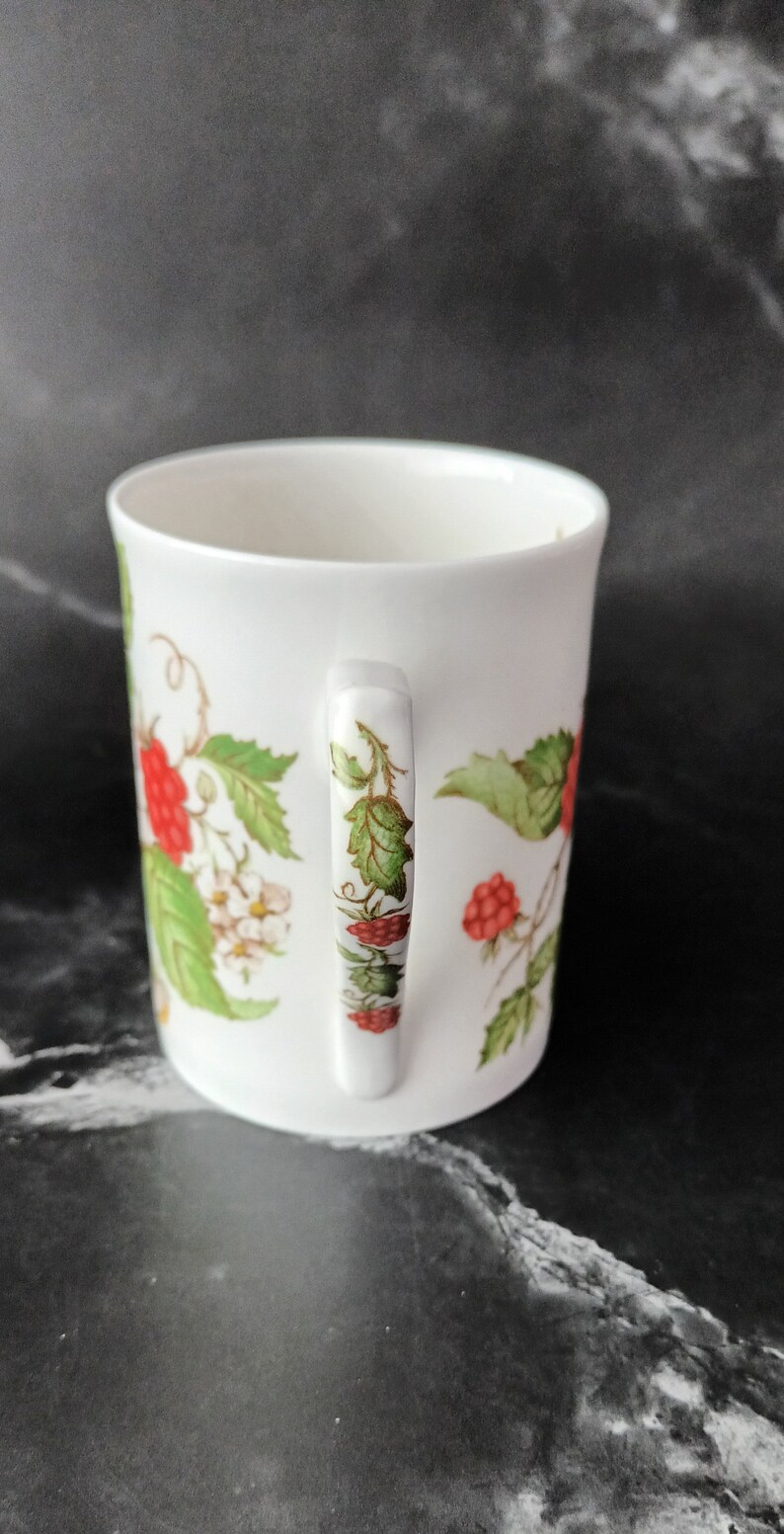 K&ouml;nnte beinhalten: Wei&szlig;e Keramiktasse mit Henkel, verziert mit einem Blumendesign mit gr&uuml;nen Bl&auml;ttern und roten Beeren.
