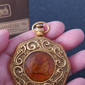 May include: Vintage gold-toned compact with ornate filigree design. The center features a reddish-brown cameo with two figures. The compact has a loop at the top and is held in a hand.