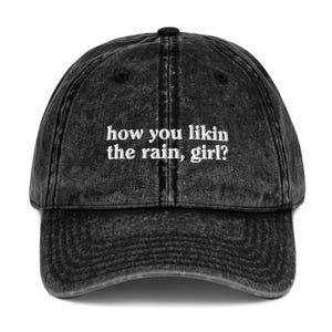 May include: Black vintage washed denim baseball cap with white embroidered text that reads "how you likin the rain, girl?"