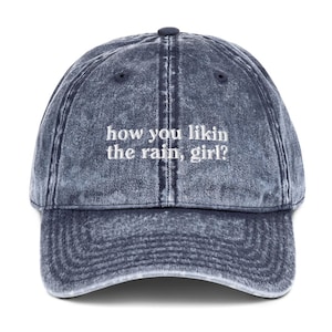 May include: A blue denim baseball cap with the text "how you likin the rain, girl?" embroidered in white.