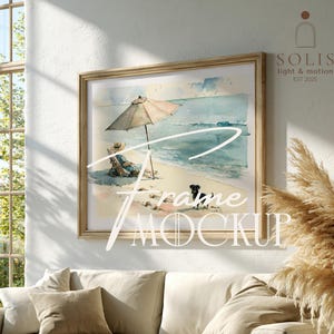 May include: A rectangular wooden frame containing a watercolor print of a beach scene with a person sitting under a tan umbrella, a small black dog on the sand, and the ocean in the background. The text "SOLIS light & motion EST 2025" is visible.