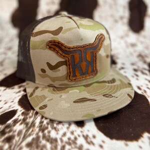 May include: A camouflage trucker hat with a brown leather patch featuring a bull's head and the initials "RX".
