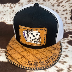 Op de afbeelding: Een zwart-witte trucker-pet met een bruine leren patch met een handgeschilderd ontwerp van speelkaarten. De pet heeft een geweven bruine leren rand met het woord "LOWMAN" in bruin garen gestikt.