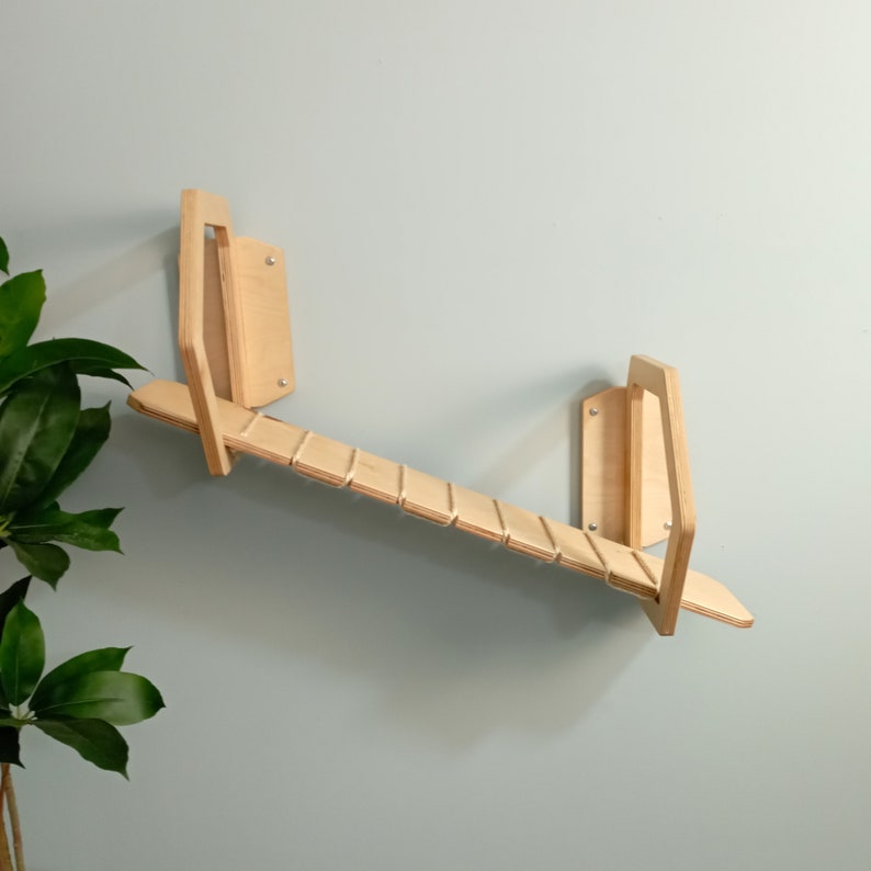 May include: A wooden wall-mounted cat climbing shelf with a ramp and two platforms. The shelf is made of natural wood and has a light brown color.