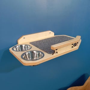 May include: A wall-mounted pet feeder with two stainless steel bowls and a gray carpet-like surface. The feeder is made of light wood and has a decorative cutout on the side.