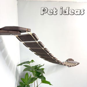 May include: A wooden cat bridge with a rope and a soft, gray fabric platform at each end. The bridge is mounted on a wall and is curved to fit a corner. The bridge is made of dark brown wood and has a natural finish. The text "Pet ideas" is visible in the upper right corner of the image.