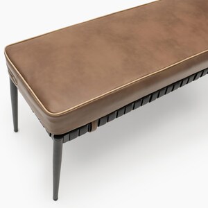 May include: A rectangular bench featuring a smooth brown upholstered seat with light tan piping. The base consists of a black frame with vertical slats and four slender, tapered black legs that extend outward from the corners of the seat.