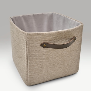 Può includere: Un contenitore quadrato con esterno in tessuto beige neutro e interno grigio chiaro. Presenta un manico in pelle marrone su un lato, fissato con rivetti color oro. Il contenitore è progettato per l'organizzazione e lo stoccaggio.