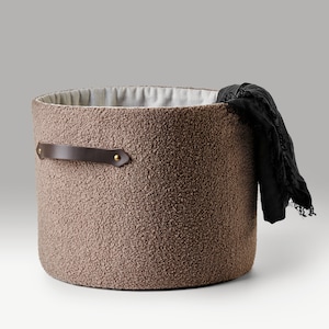 May include: A brown fuzzy storage basket with a leather handle and a black fabric item inside.