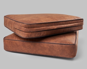 May include: Two square, brown cushions stacked. They have a textured appearance and are trimmed with dark brown piping. The cushions appear to be made of a leather-like material. The cushions are suitable for indoor or outdoor use.
