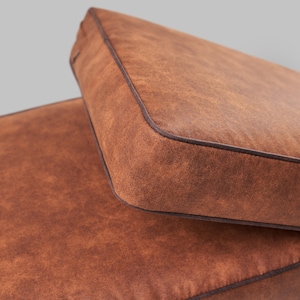 Może przedstawiać: Zbliżenie dwóch brązowych poduszek z ciemnobrązową lamówką. Poduszki mają teksturowany wygląd przypominający skórę. Poduszki są prostokątne z zaokrąglonymi rogami i prawdopodobnie przeznaczone do mebli ogrodowych.