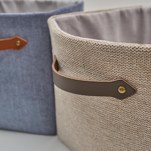Può includere: Due contenitori portaoggetti in tessuto. Uno è in un materiale blu chiaro simile al denim, e l'altro è in tessuto beige intrecciato. Entrambi hanno manici in pelle marrone fissati con rivetti in ottone. Gli interni sono foderati con tessuto grigio chiaro.