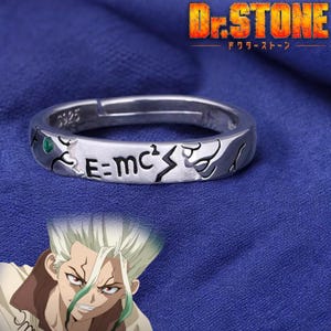 Puede incluir: Anillo de plata con una piedra verde y la inscripción "E=mc²" y un rayo. El anillo es de la serie de anime Dr. Stone, que está escrito en japonés en el anillo.