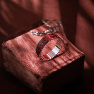 May include: A silver ring with a red and silver design. The ring is on a wooden surface.