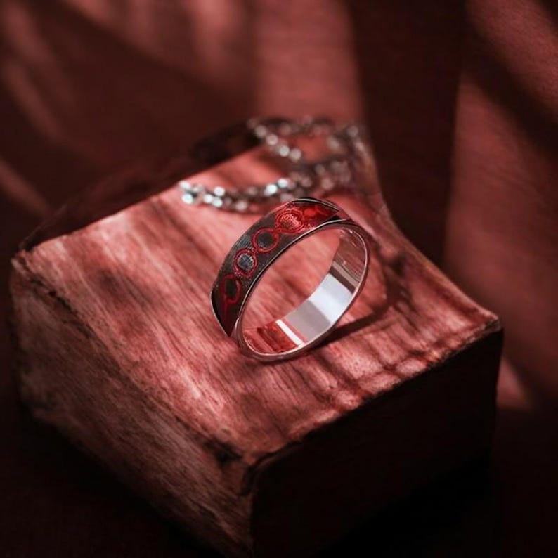 May include: A silver ring with a red and silver design. The ring is on a wooden surface.