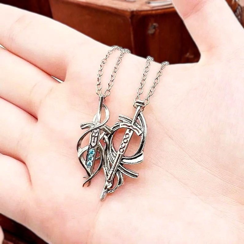 May include: Two silver necklaces with a sword and a dagger pendant. The pendants are intertwined and feature blue gemstones.