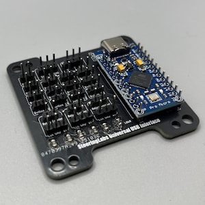 Op de afbeelding: Een zwart SteeringLabs Universal USB Interface bord met een blauw Pro Micro bord bevestigd. Het zwarte bord heeft meerdere pinnen en labels zoals "Button" en "Rotary Encoder". Het blauwe bord heeft een USB-C poort en diverse elektronische componenten.