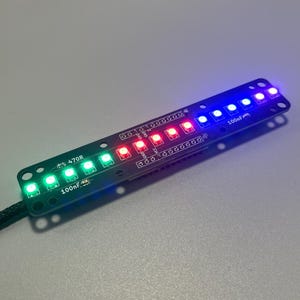 Op de afbeelding: Een lange, smalle zwarte printplaat met een rij van tien opbouw-LED's die oplichten in een verloop van groen, rood en blauw. De printplaat bevat witte bedrukte labels zoals 470R en 100nF, samen met diverse pin-headers voor aansluitingen.