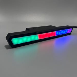 Peut inclure: Une barre lumineuse LED noire avec des sections de lumières vertes, rouges et bleues. La barre lumineuse a une finition mate et un câble. Le texte "STEALTH LABS" est visible sur le dessus. Mesure environ 20 cm de long.