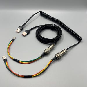 Könnte beinhalten: Schwarzes, gewendeltes USB-Kabel mit einem USB-A-Anschluss und einem USB-C-Anschluss. Das Kabel hat einen gewendelten Abschnitt und zwei gerade Abschnitte mit silberfarbenen Anschlüssen. Die Drähte sind mehrfarbig.