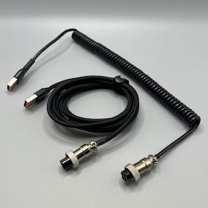Könnte beinhalten: Schwarzes, gewendeltes Tastaturkabel mit USB-A- und USB-C-Anschlüssen. Das Kabel hat einen gewendelten und einen geraden Abschnitt mit silberfarbenen Anschlüssen an jedem Ende. Das Kabel ist für mechanische Tastaturen konzipiert.