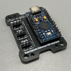 Op de afbeelding: Een zwarte printplaat met het label SteeringLabs Universal USB Interface en een blauwe Pro Micro microcontroller bevestigd. De kaart bevat drie sets pinnen voor Accelerator, Brake en Clutch, elk met 5V, GND en Analog Signal labels.