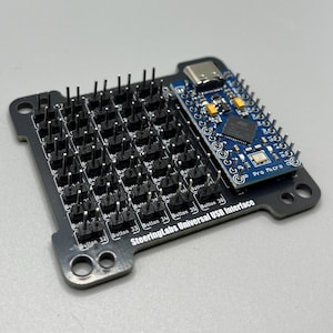 Könnte beinhalten: Ein schwarzes SteeringLabs Universal USB Interface mit zahlreichen Button-Pins und einem blauen Pro Micro Board. Die Schnittstelle verfügt über beschriftete Tasten und einen USB-C-Anschluss, konzipiert für Elektronikprojekte.