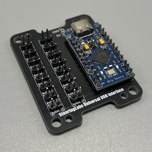 Könnte beinhalten: Eine schwarze Leiterplatte mit der Aufschrift SteeringLabs Universal USB Interface, zwei Reihen von Stiftleisten und einem blauen Pro Micro Mikrocontroller-Modul. Die Platine weist weiße Textmarkierungen für die Tasten 1 bis 16 sowie GND/5V auf.