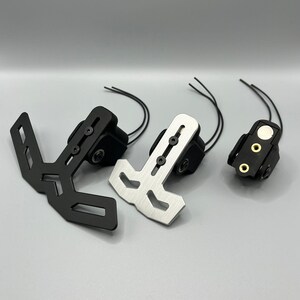 Pode incluir: Três suportes de placa de matrícula de motocicleta em preto e prata. Os suportes têm várias formas e designs, incluindo uma forma de T e um suporte com secções recortadas. Cada suporte está preso a uma caixa preta com fios que se estendem dela.