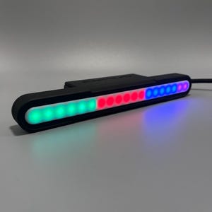 Peut inclure: Une barre lumineuse LED noire avec un design incurvé. La barre affiche un dégradé de couleurs, passant du vert au rouge puis au bleu. La barre lumineuse est sur une surface grise.