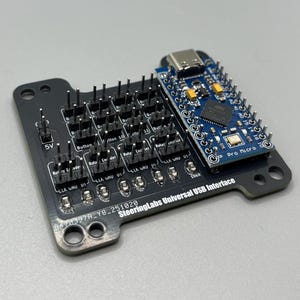 Puede incluir: Una placa de circuito negro SteeringLabs Universal USB Interface con una placa azul Pro Micro adjunta. La placa negra tiene múltiples pines, secciones etiquetadas y el texto "5V". La placa azul tiene un puerto USB y varios pines.