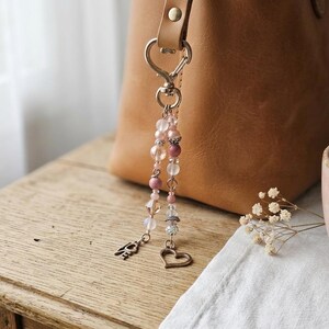 May include: A decorative bag charm featuring two dangling strands of beads in shades of pink, clear, and white. The charm includes a silver-toned metal heart-shaped clasp, a small metal heart charm, and a metal charm with the word LOVE attached.
