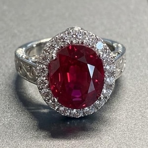 Puede incluir: Un anillo de tono plateado con una gran piedra preciosa de color rojo intenso y corte ovalado en el centro. La piedra central está rodeada por un halo de pequeñas piedras claras y brillantes. La banda del anillo muestra un patrón de filigrana.