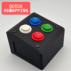 May include: A black square device featuring four coloured buttons: blue, green, red, and white. The text "QUICK REMAPPING" is in a red rectangle. The device has a textured surface and is secured with black screws.