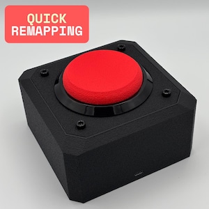 Könnte beinhalten: Ein schwarzer quadratischer Knopf mit einem großen roten Knopf oben. Der Knopf ist von einem schwarzen Ring umgeben und mit vier Schrauben befestigt. Der Text "QUICK REMAPPING" befindet sich in einem roten Rechteck oben links.