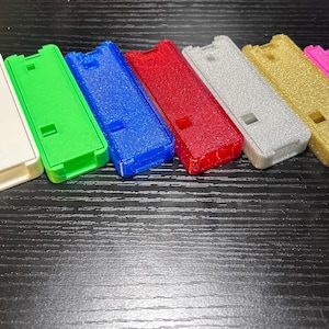 May include: A collection of eight rectangular, plastic cases in various colors, including black, white, green, blue, red, silver, gold, and pink. Each case has a rectangular opening and a small square cutout.