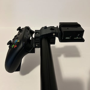 May include: A black video game controller and a black box with a cable attached are mounted to a black pole.