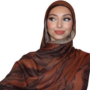 May include: A brown and black patterned hijab. The fabric drapes over the head and shoulders, creating a soft, flowing effect. The design features a marbled pattern in shades of brown and black.