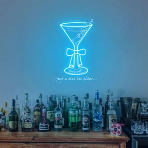 May include: A neon sign of a martini glass with a blue glow and the text "just a tiny bit older..."  The sign is hanging on a wall above a wooden cabinet with a variety of liquor bottles.