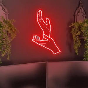 Può includere: Un'insegna al neon rossa raffigurante due mani che si tendono l'una verso l'altra. L'insegna è montata su una parete scura, con piante sospese e due monitor per computer sottostanti. L'estetica generale è moderna e artistica.
