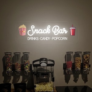 May include: A snack bar setup with a neon sign that reads "Snack Bar Drinks - Candy - Popcorn". There are three dispensers filled with candy, a popcorn machine, and two buckets.
