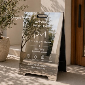 Pode incluir: Uma placa de calçada em forma de A de tom prateado com uma superfície reflexiva. O texto diz: Welcome! We're Open, PETERSON MADE, Open Today Come In & Explore, Fine Jewelry, Custom Design, Gifts, Repairs, Celebrate Life's Moments, Wear Your Story.